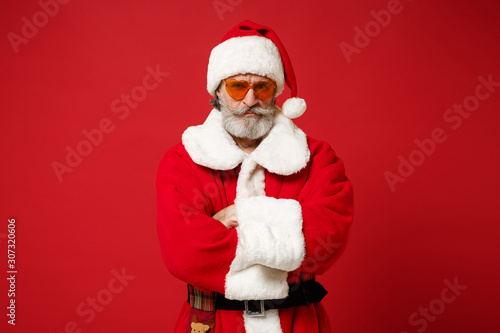 Serious Elderly gray-haired mustache bearded Santa man in Christmas hat, sunglasses posing isolated on red background. Happy New Year 2020 celebration concept. Mock up copy space. Hold hands crossed.
