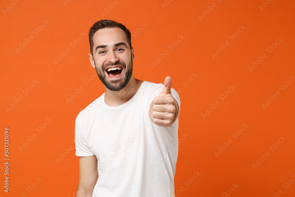 Excited funny young man in casual white t-shirt posing isolated on bright orange wall background, studio portrait. People sincere emotions lifestyle concept. Mock up copy space. Showing thumb up. Excited funny young man in casual white t-shirt posing isolated on bright orange wall background, studio portrait. People sincere emotions lifestyle concept. Mock up copy space. Showing thumb up.