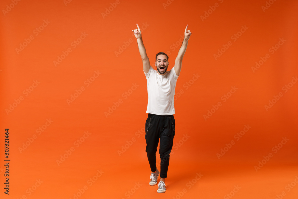 Funny young man in casual white t-shirt posing isolated on orange background, studio portrait. People sincere emotions lifestyle concept. Mock up copy space. Rising hands, pointing index fingers up. - obrazy, fototapety, plakaty 