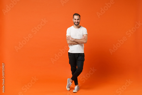 Smiling attractive young man in casual white t-shirt posing isolated on orange wall background studio portrait. People sincere emotions lifestyle concept. Mock up copy space. Holding hands crossed.
