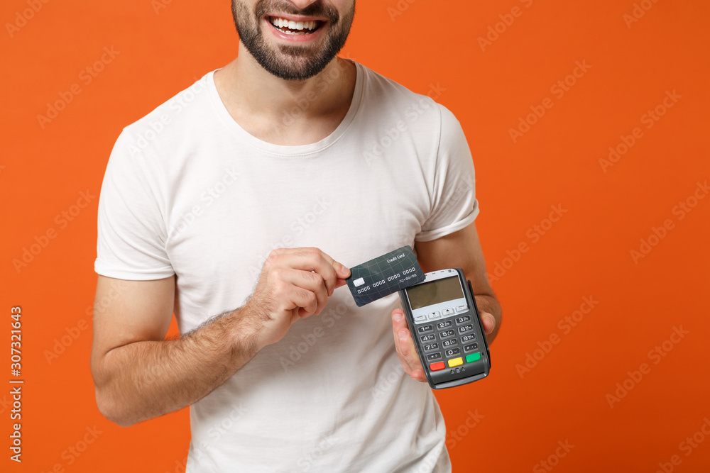 Cropped image of man in white t-shirt posing isolated on orange background. People lifestyle concept. Mock up copy space. Hold wireless bank payment terminal to process acquire credit card payments. Cropped image of man in white t-shirt posing isolated on orange background. People lifestyle concept. Mock up copy space. Hold wireless bank payment terminal to process acquire credit card payments.