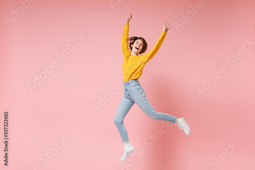 Cheerful young brunette woman girl in yellow sweater posing isolated on pastel pink background in studio. People lifestyle concept. Mock up copy space. Having fun fooling around rising hands, jumping.