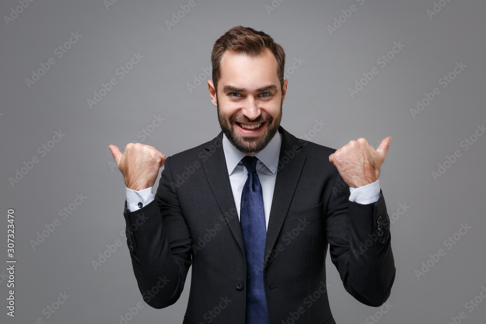 Cheerful young bearded business man in classic black suit shirt tie posing isolated on grey background in studio. Achievement career wealth business concept. Mock up copy space. Pointing thumbs aside. Cheerful young bearded business man in classic black suit shirt tie posing isolated on grey background in studio. Achievement career wealth business concept. Mock up copy space. Pointing thumbs aside.