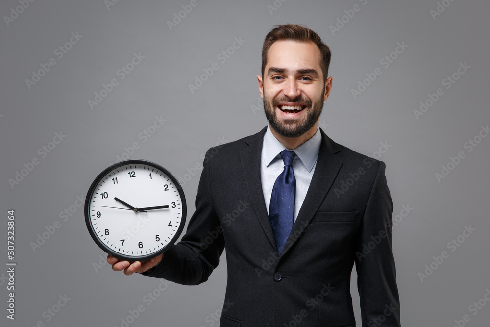 Laughing young bearded business man in classic black suit shirt tie posing isolated on grey background studio portrait. Achievement career wealth business concept. Mock up copy space. Holding clock. Laughing young bearded business man in classic black suit shirt tie posing isolated on grey background studio portrait. Achievement career wealth business concept. Mock up copy space. Holding clock.