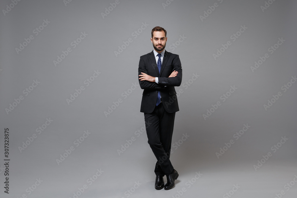 Handsome young bearded business man in classic black suit shirt tie posing isolated on grey background in studio. Achievement career wealth business concept. Mock up copy space. Holding hands crossed. - obrazy, fototapety, plakaty 