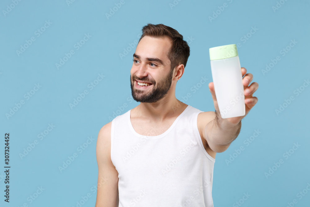 Bearded young man 20s years old in white shirt hold in hand bottle tonic isolated on blue pastel wall background, studio portrait. Skin care healthcare cosmetic procedures concept. Mock up copy space. Bearded young man 20s years old in white shirt hold in hand bottle tonic isolated on blue pastel wall background, studio portrait. Skin care healthcare cosmetic procedures concept. Mock up copy space.