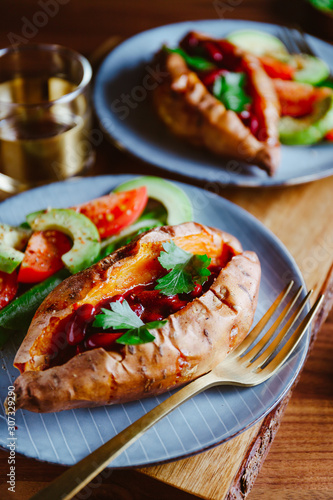 Baked sweet potato filled with red beans stewed in tomato paste served with avocado and cherry tomatoes on a plate. Healthy veggie recipe.