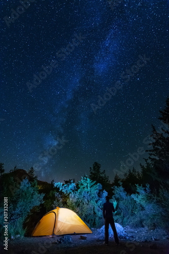 Vertical landscape images with a night starry sky and a milky way against the backdrop of a yellow tourist tent, which is highlighted from the inside.