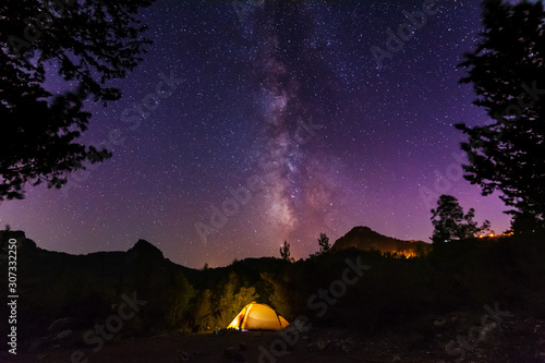 Vertical landscape images with a night starry sky and a milky way against the backdrop of a yellow tourist tent, which is highlighted from the inside.