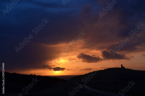 Hügelige Landschaft Silhouette mit Leuchtturm bei Sonnenuntergang vor bewölktem Himmel
