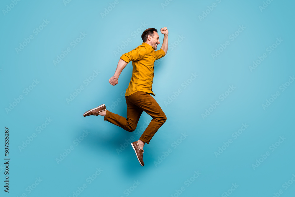 Full size profile side photo of cheerful positive man jump run after spring time sales wear casual style outfit sneakers isolated over blue color background Full size profile side photo of cheerful positive man jump run after spring time sales wear casual style outfit sneakers isolated over blue color background