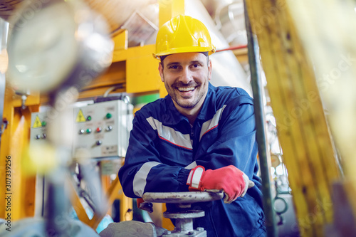 Photo Handsome caucasian smiling unshaven worker in protective uniform leaning on valve while standing in factory