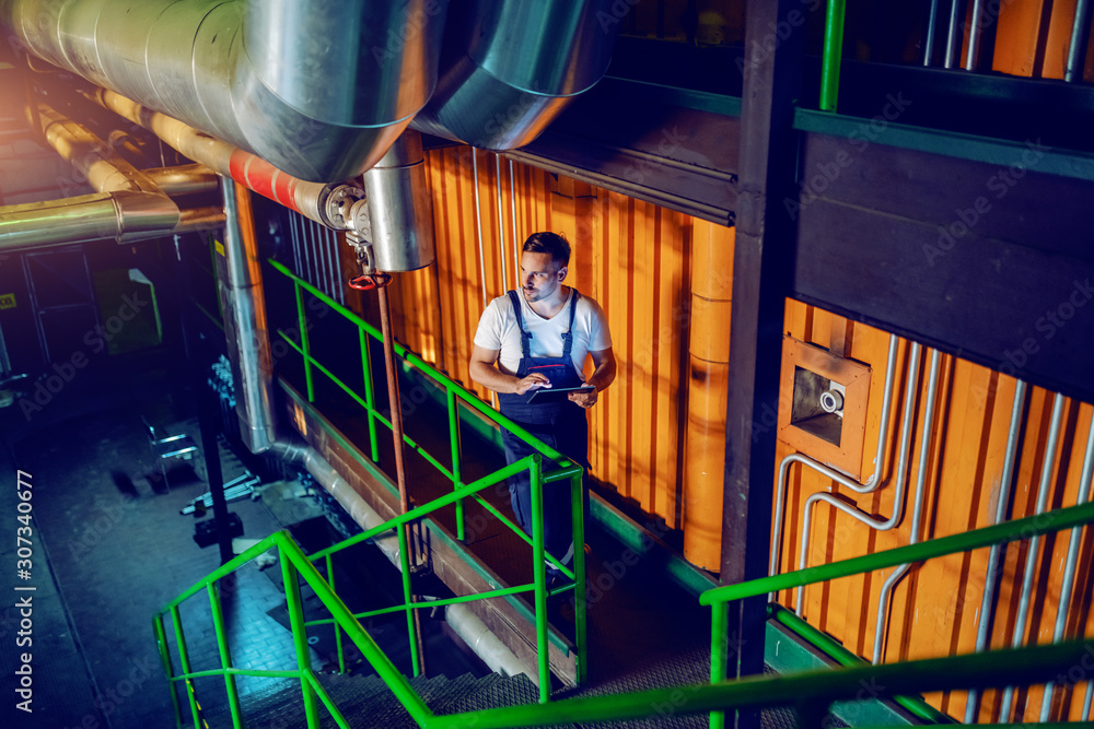Full length of serious worker in overall holding tablet and walking around factory. Full length of serious worker in overall holding tablet and walking around factory.