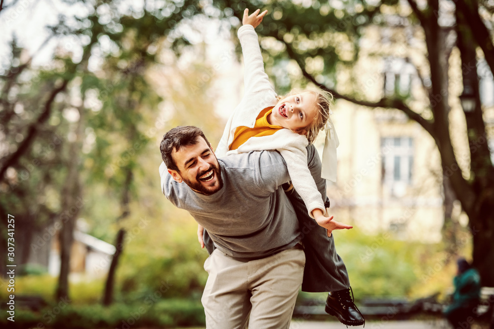 Playful handsome bearded Caucasian father having piggyback with his adorable daughter. Autumn in park.