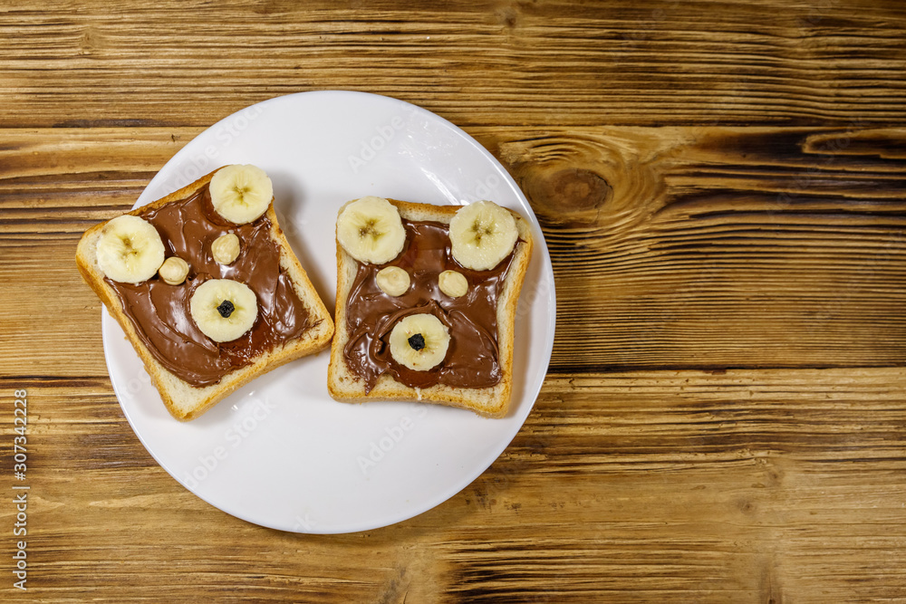 Two sweet sandwiches with delicious chocolate hazelnut spread and banana in shape of bear on wooden table. Top view Two sweet sandwiches with delicious chocolate hazelnut spread and banana in shape of bear on wooden table. Top view