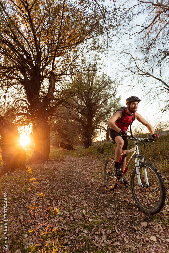 Happy determined young bearded man in red sleeveless cycling jersey riding mountain bike through the forest during sunset. Sun setting between trees in the background.