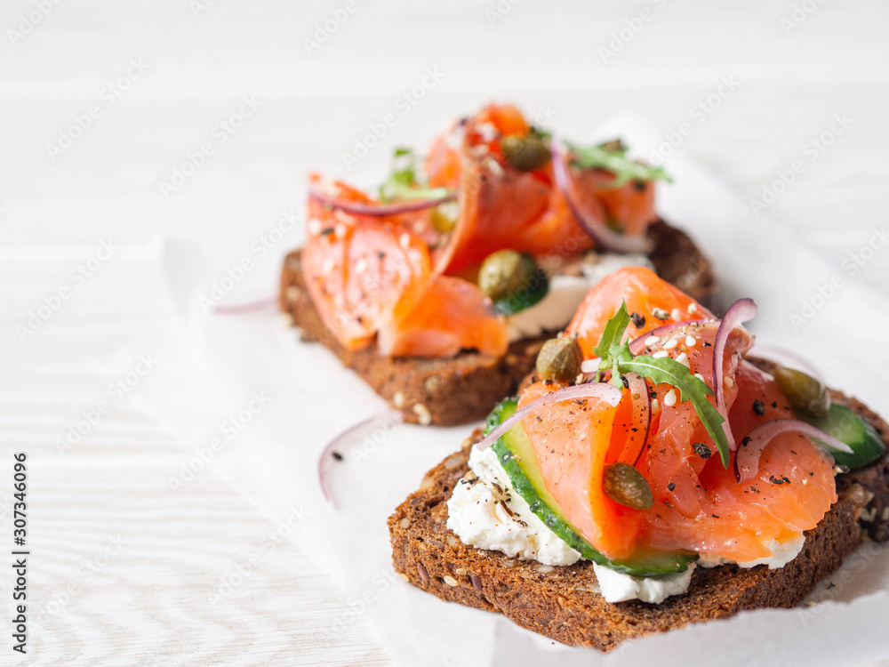 Healthy toasts with rye bread with cream cheese, salmon, fresh cucumber, capers, sesame seeds, black pepper and arugula on white paper. Copy space