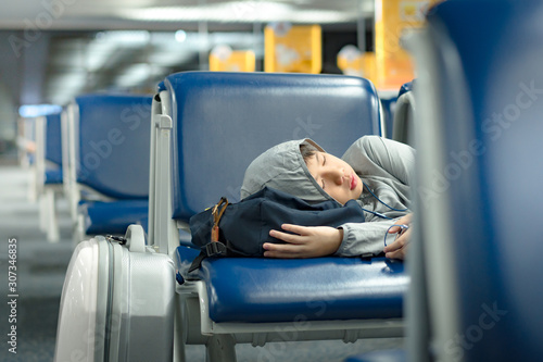 Young Asian teenage boy, solo traveler sleep on seat in modern airport terminal, listen to music.
