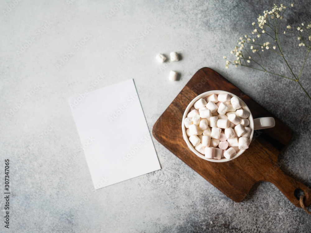 White cup with hot drink and marshmallows and flowers and white card on a grey background. Copy space. Top view White cup with hot drink and marshmallows and flowers and white card on a grey background. Copy space. Top view