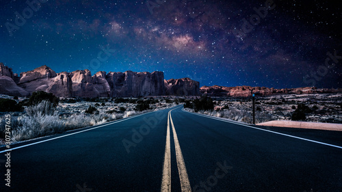 An endless desolate road leading into Arches National Park in Moab, Utah, USA under a dark and starry night sky.