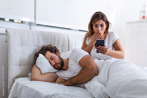 Fotografie Jealous wife spying the phone of her partner while he is sleeping in a bed at home