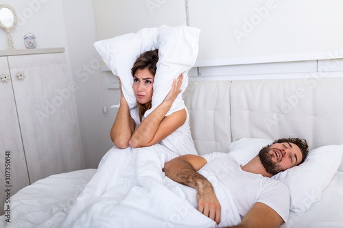 Young couple have problem with man's snoring. Heterosexual couple in bed, man sleeps and snoring with mouth open, while a tired woman irritated by snoring sitting on bed with a pillow on her head.