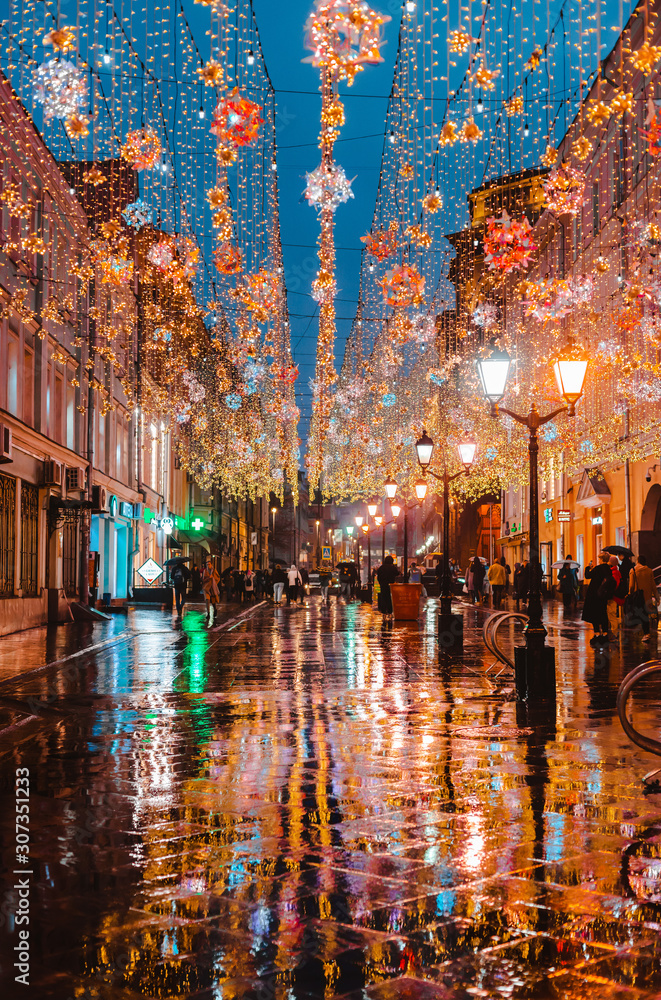 rainy night in a big city, the reflection of colorful city lights on the wet road surface. View of a pedestrian street with bright city holiday illumination.