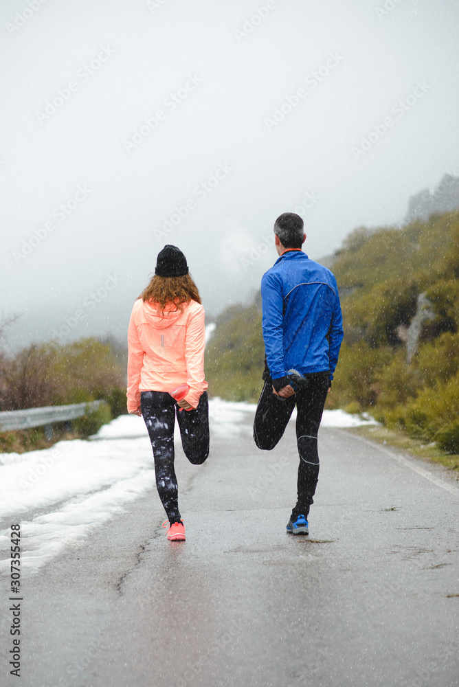 Back view of sporty couple of runners stretching legs for warming up before running in winter mountain road under the snow. Back view of sporty couple of runners stretching legs for warming up before running in winter mountain road under the snow.