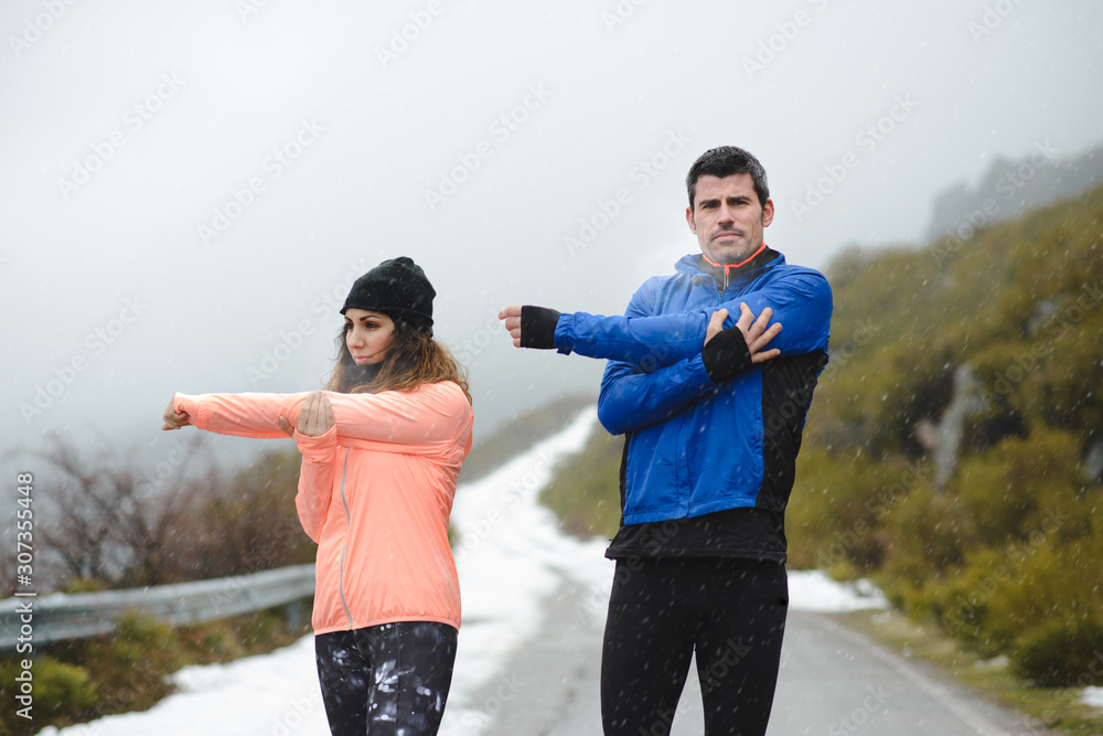 Couple of runners stretching arms and shoulders for warming up before running in winter mountain road under the snow. Couple of runners stretching arms and shoulders for warming up before running in winter mountain road under the snow.