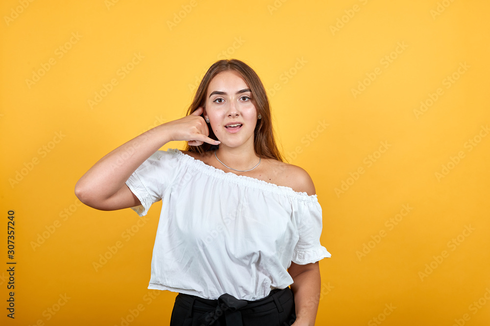 Attractive young woman doing telephone gesture, looking shocked isolated on orange background in studio in casual white shirt. People sincere emotions, lifestyle concept. Attractive young woman doing telephone gesture, looking shocked isolated on orange background in studio in casual white shirt. People sincere emotions, lifestyle concept.