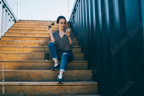 Joyful adult woman with hot drink to go using smartphone on stairs in city