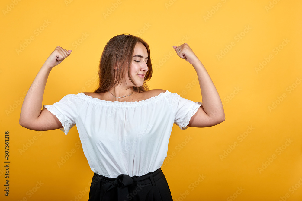 Strong young woman showing muscles, looking happy isolated on orange background in studio in casual white shirt. People sincere emotions, lifestyle concept. Strong young woman showing muscles, looking happy isolated on orange background in studio in casual white shirt. People sincere emotions, lifestyle concept.