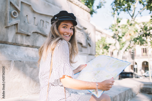 Portrait of cheerful Caucasian female tourist with paper map in hands smiling at camera during researching location of beautiful showplaces in Spanish city - Barcelona, time for travel to Europe #307362026