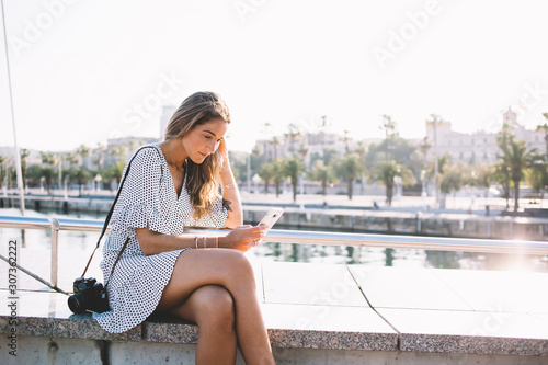 Attractive female photographer taking rest in port while sending sms message and waiting model for photography in Barcelona port, beautiful hipster girl reading information on website via gadget © BullRun