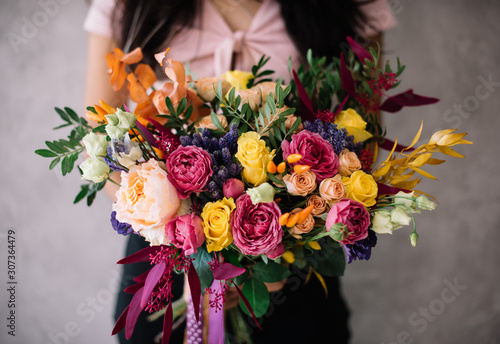 Very nice young woman holding big blossoming bouquet of campanella peach roses, misty bubbles roses, ranunculus, eustoma, pistachio fresh flowers in pink and yellow colors on the grey wall background photo