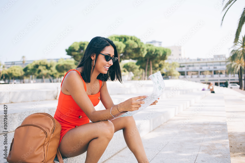 Latin brunette hipster girl taking rest on urban setting in Spanish city enjoying time for international vacations, carefree woman checking information from paper location map and smiling outdoors
