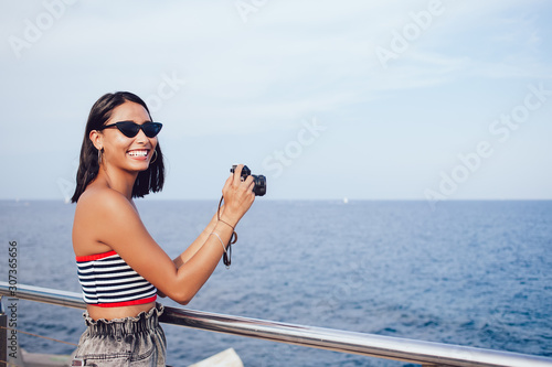 Portrait of toothy female amateur photographer in trendy fox sunglasses smiling at camera during time for taking pictures of seashore landmark using modern SLR technology during summer promenade