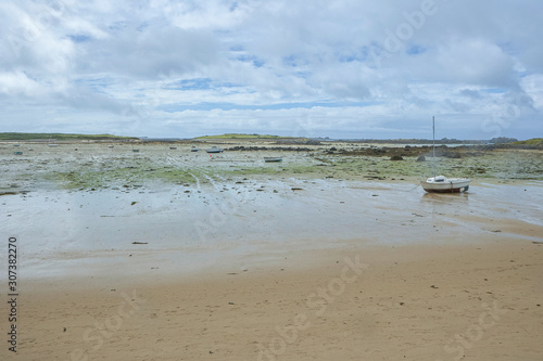 Coast in Brittany at low tide with sand and seaweed and small boats, cloudy blue sky, silent mood