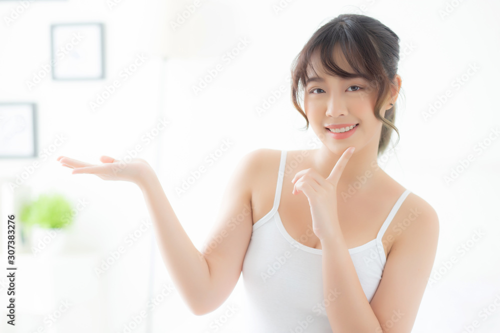 Beautiful young asian woman smiling and showing skin care product for advertisement at bedroom, beauty asia girl presenting makeup cosmetic for advertise with surprise on palms at room. Beautiful young asian woman smiling and showing skin care product for advertisement at bedroom, beauty asia girl presenting makeup cosmetic for advertise with surprise on palms at room.