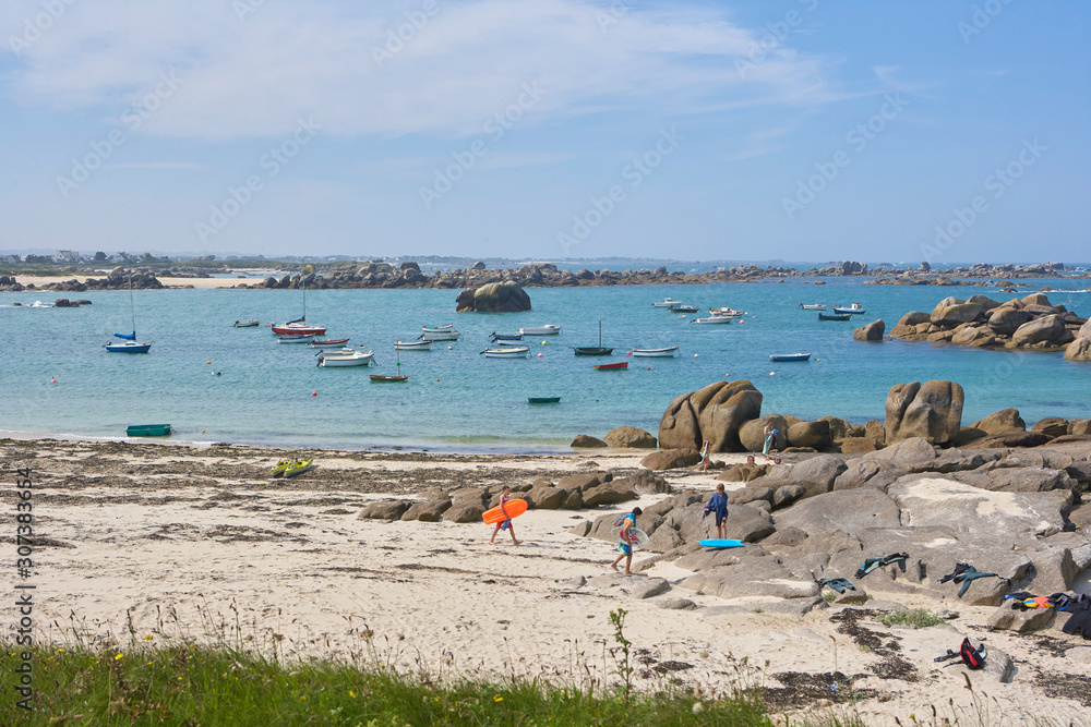 Small beach with tourists with surfboards, round rocks, sand, little boats in the calm sea, summer holiday feeling