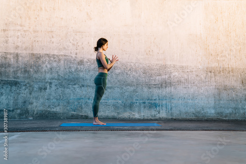 Young serene brunette making Prayer hands yoga pose on fitness mat against concrete wall