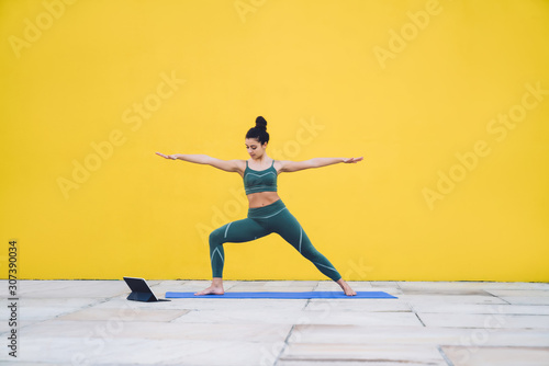 Young sporty slim woman exercising yoga in Virabhadrasana II pose using tablet in yard