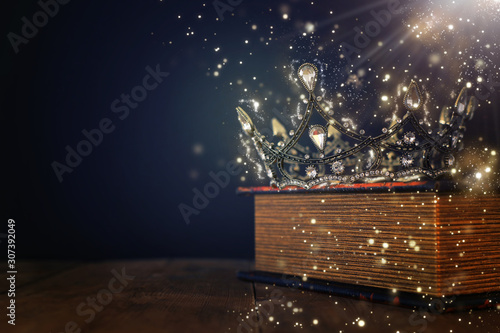 low key image of beautiful queen/king crown over old book and wooden table. vintage filtered. fantasy medieval period. Glitter sparkle lights