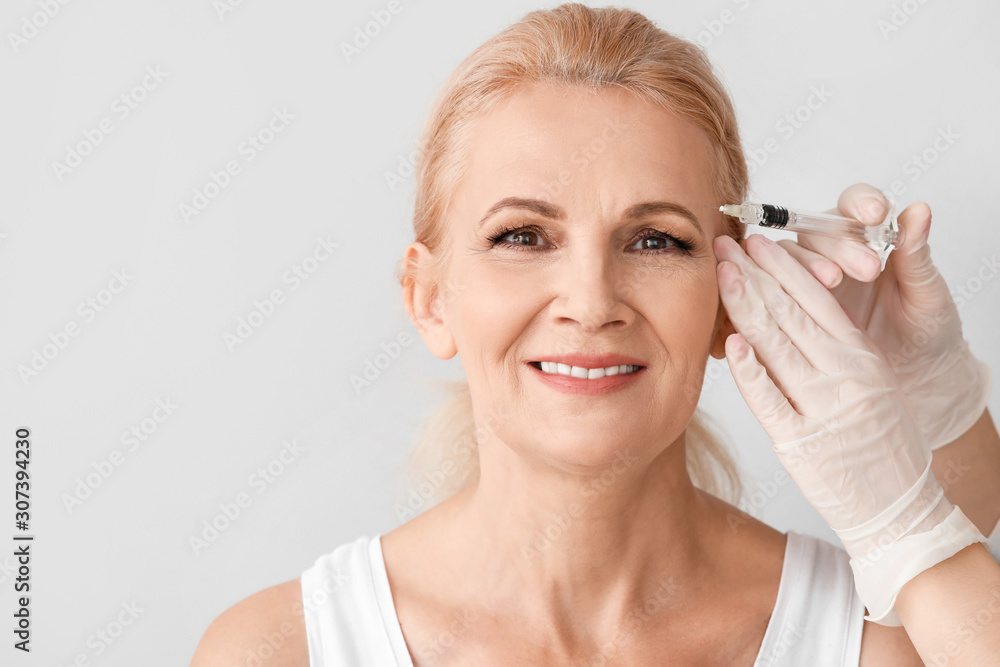 Fototapeta premium Mature woman receiving anti-aging injection in her face against light background