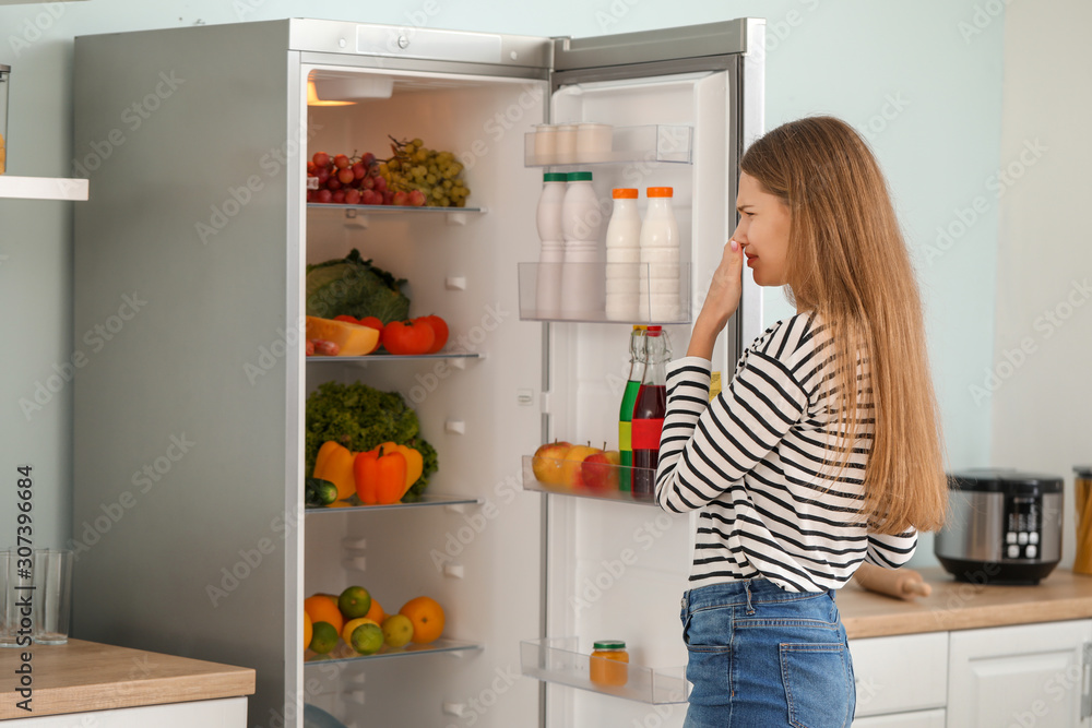 Woman feeling bad smell from fridge in kitchen - obrazy, fototapety, plakaty 