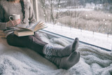 Woman sitting by the window reading book drinking coffee. Winter snowing landscape outside 