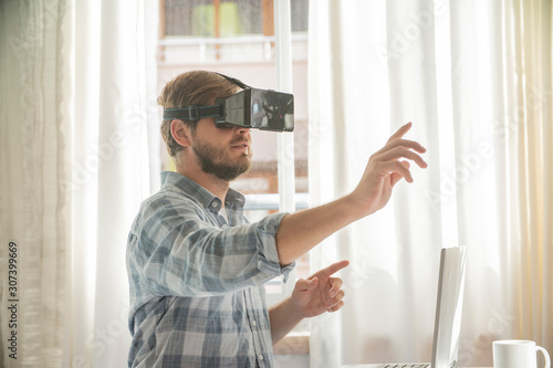 Young bearded man wearing virtual reality glasses in modern interior. Handsome caucasian male using VR headset, playing video games.