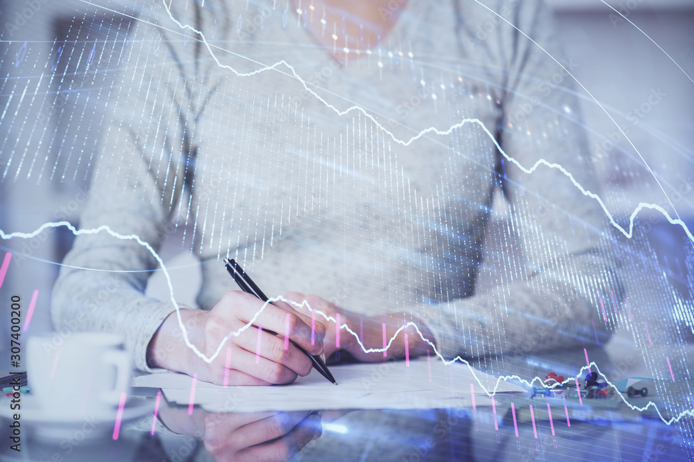 Fototapeta premium Hand taking notes in notepad. Forex chart holograms in front. Concept of research. Multi exposure
