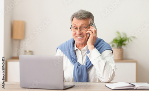 Elderly Man Talking On Phone Working On Laptop At Workplace