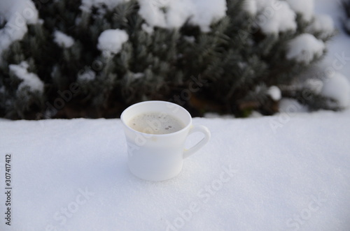Winter. December. Against the backdrop of a snowy lavender bush, there is a white cup of freshly brewed aromatic coffee.